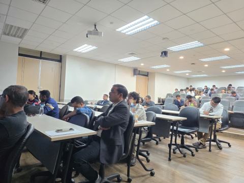 Attendees seated at desks in the seminar room follow a talk, with more participants in the tiered seats behind them.
