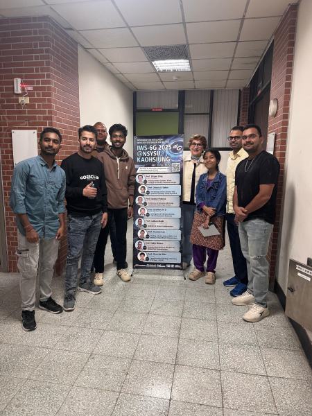 Eight participants and lab members stand on both sides of the IWS-6G 2025 @NSYSU, Kaohsiung roll-up banner, which lists the invited speakers and their talks, in a corridor of the workshop building.