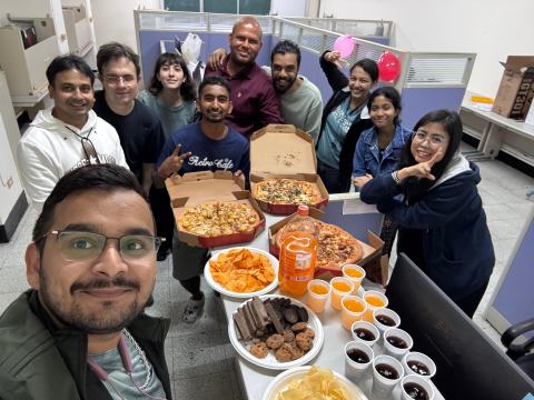 Selfie of ten lab members around a table with pizzas, crisps, cookies and drinks in the lab office, with balloons on the cubicle partitions.